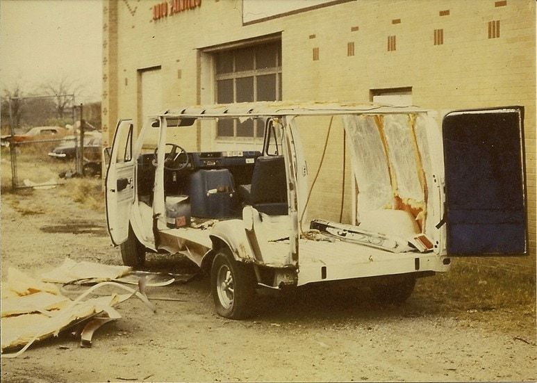 An old photo of a white van with its side torn open, parked beside the GAC shop building